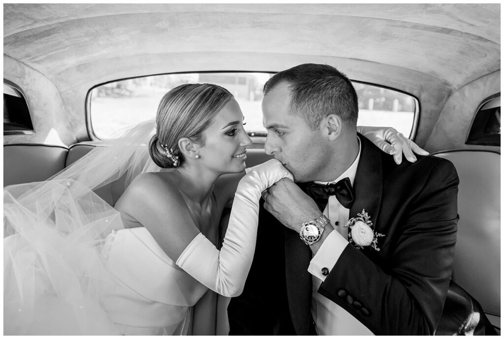 Groom leaning in to kiss the hand of his elegant bride in the back of a vintage Rolls Royce. 