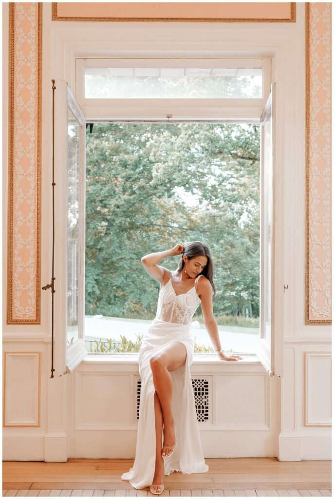 Brunette bride in lace white dress sitting, posing by a window in pink ballroom at Cairnwood Estate. 
