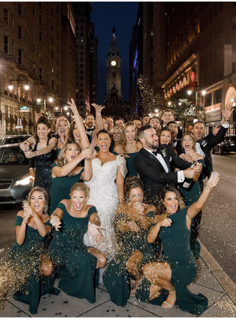 Wedding party surround the bride and groom as they pop confetti outside of The Lucy with a beautiful view of Philadelphia City Hall at night behind them