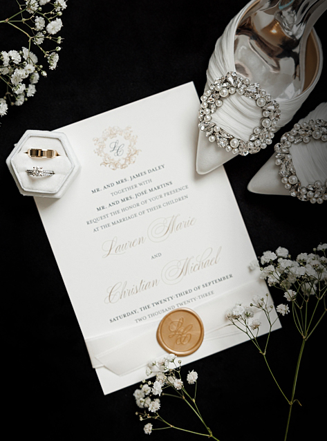 wedding flat lay of white and gold invitations laying on a black table with small white flowers, a white velvet ring box and white wedding heels